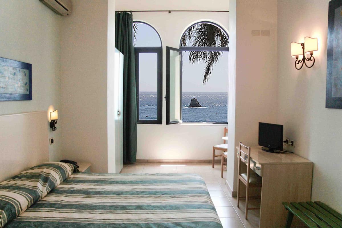 Une chambre avec un lit, un bureau, une chaise, une télévision et une fenêtre donnant sur la mer.