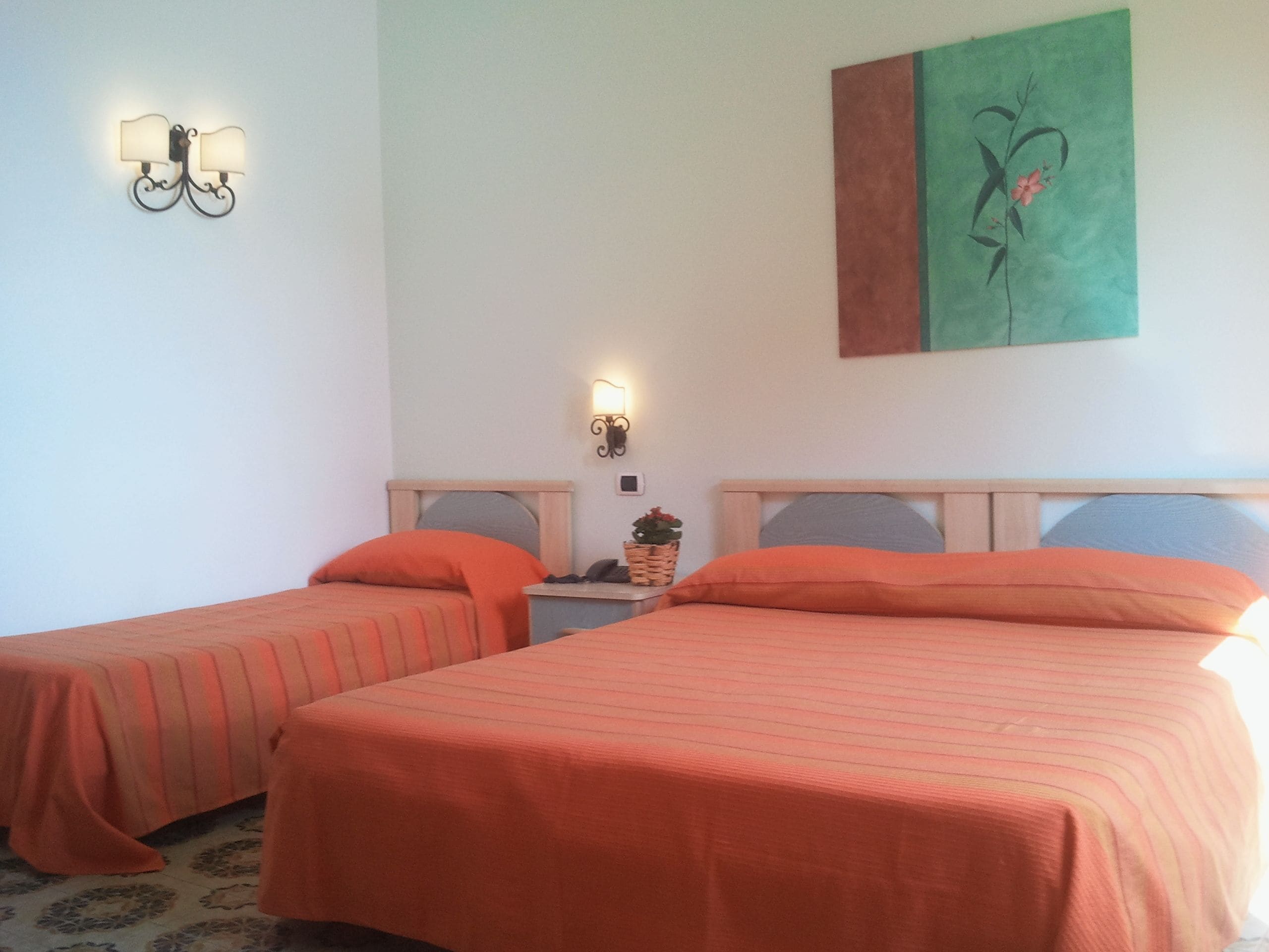 A hotel room with two beds, each with an orange bedspread, a nightstand with a lamp, and a wall-mounted artwork.