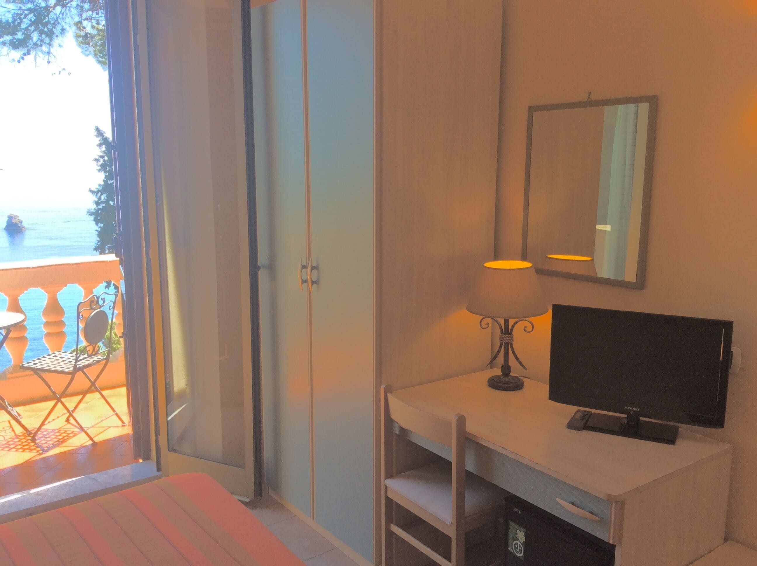 Quarto com cama, escrivaninha com computador, cadeira, luminária, televisão e varanda com cadeira e mesa.