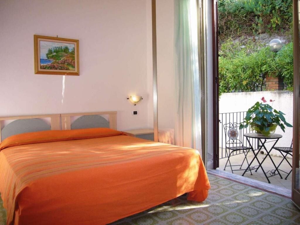 A hotel room with a bed, a painting on the wall, and a balcony with a table and chairs.