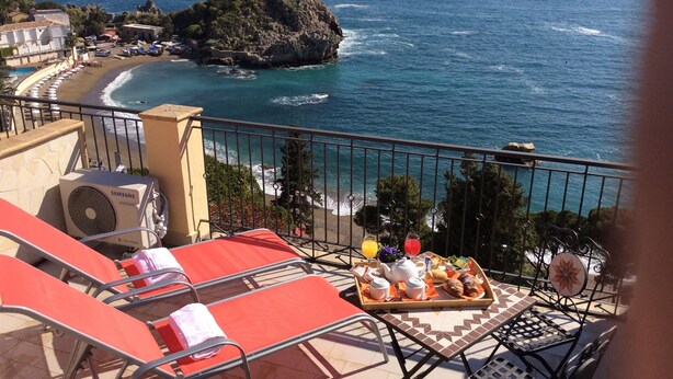 Un balcon avec des fauteuils de relaxation rouges, un plateau contenant des éléments pour le petit-déjeuner et une vue sur la mer et une île rocheuse.