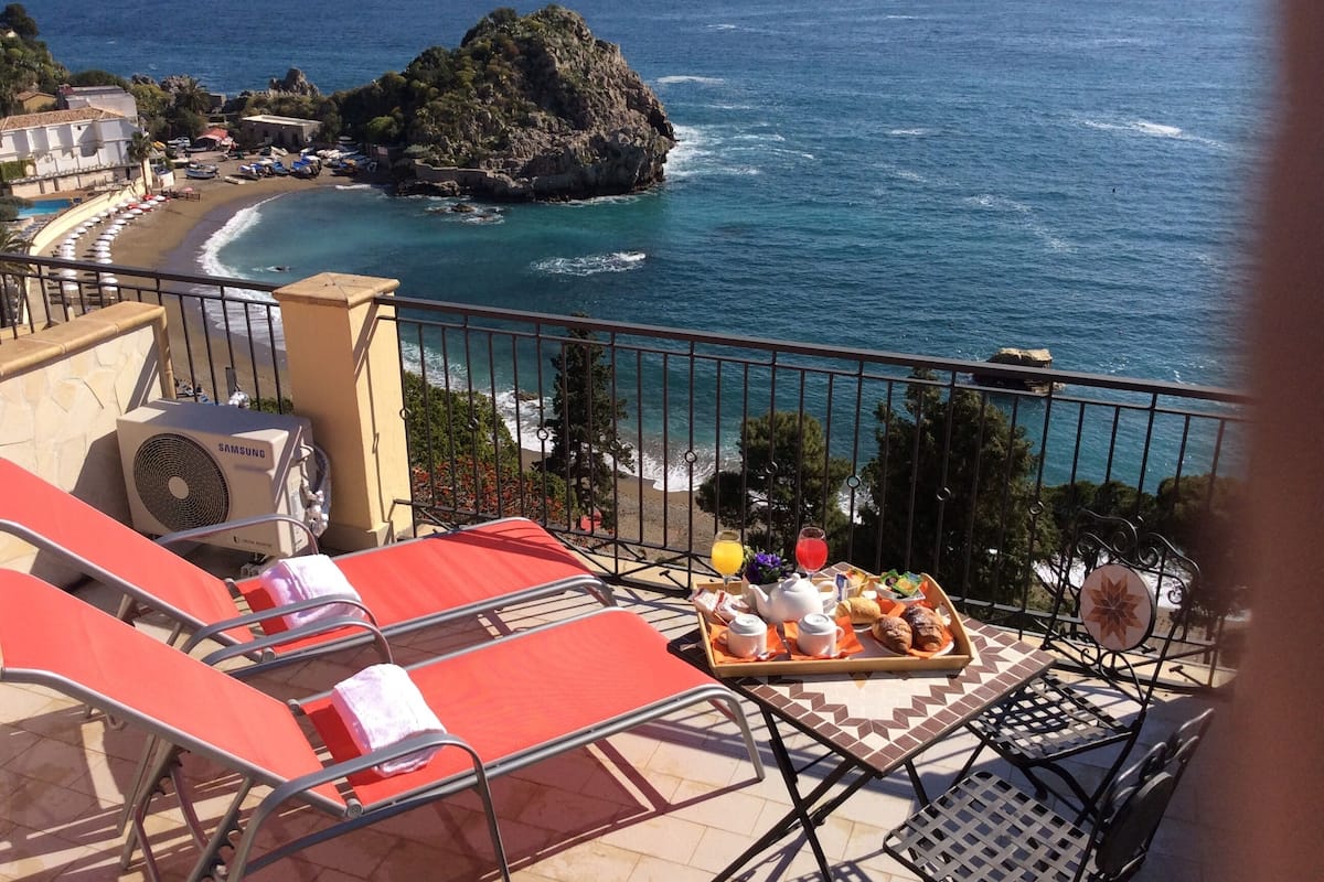 Un balcon avec des fauteuils rouges, un plateau de petit-déjeuner et une vue sur la mer et une île rocheuse.