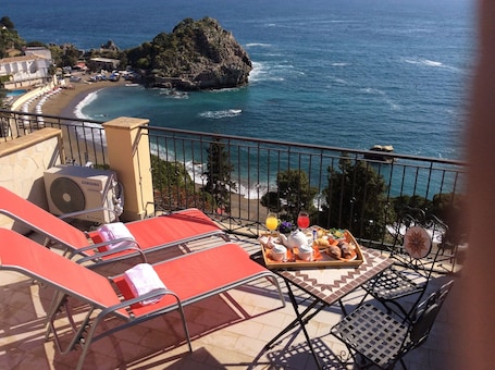 A balcony with red lounge chairs, a tray of breakfast, and a view of the sea and a rocky island.