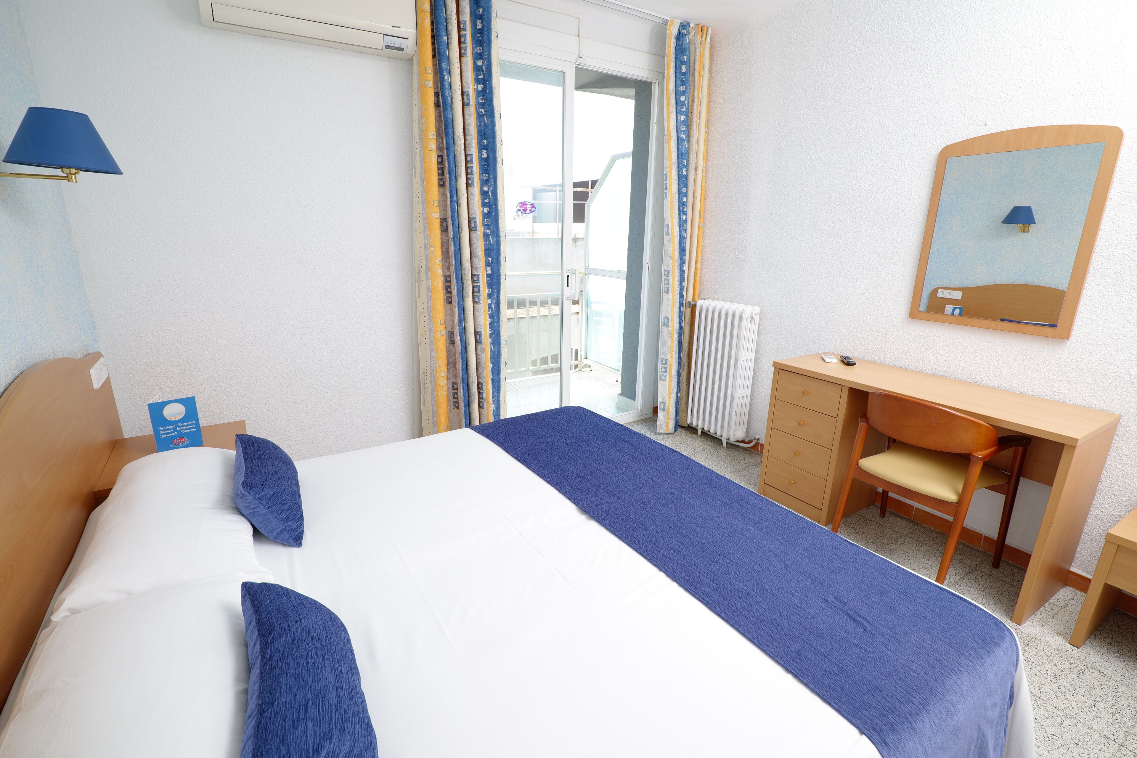 A hotel room with a bed, a desk, a chair, a lamp, and a window with curtains.
