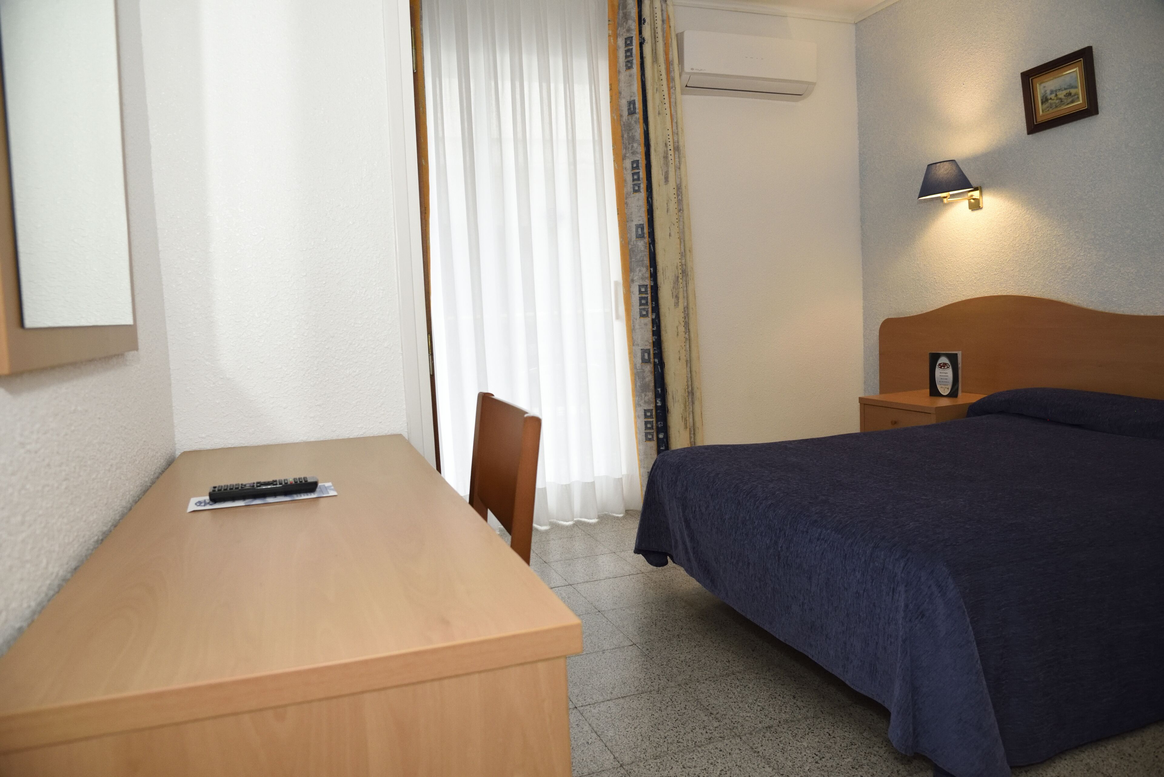 Una habitación de hotel con un escritorio de madera, una cama con cubrecama azul, una mesita de noche con lámpara y una ventana con cortinas.