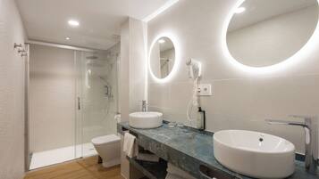 Un baño moderno con cabina de ducha de vidrio, inodoro blanco, dos lavamanos con espejos redondos y encimera de mármol.