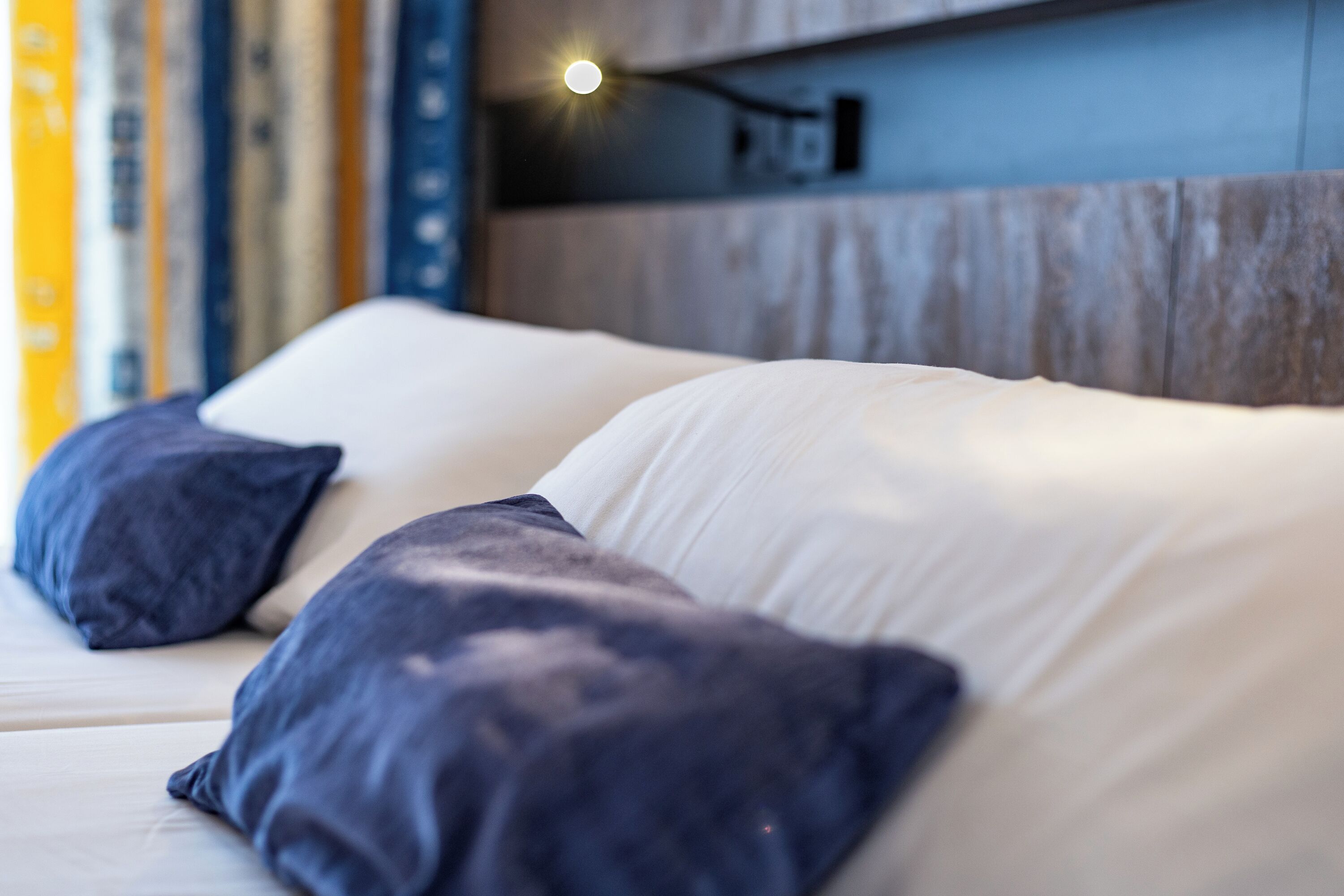 A bed with white and dark blue pillows, a wooden headboard, and a wall-mounted light fixture.