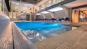 Piscina com cadeiras azuis e guarda-corpo de vidro, cercada por um edifício moderno com fachada de vidro e escadaria.