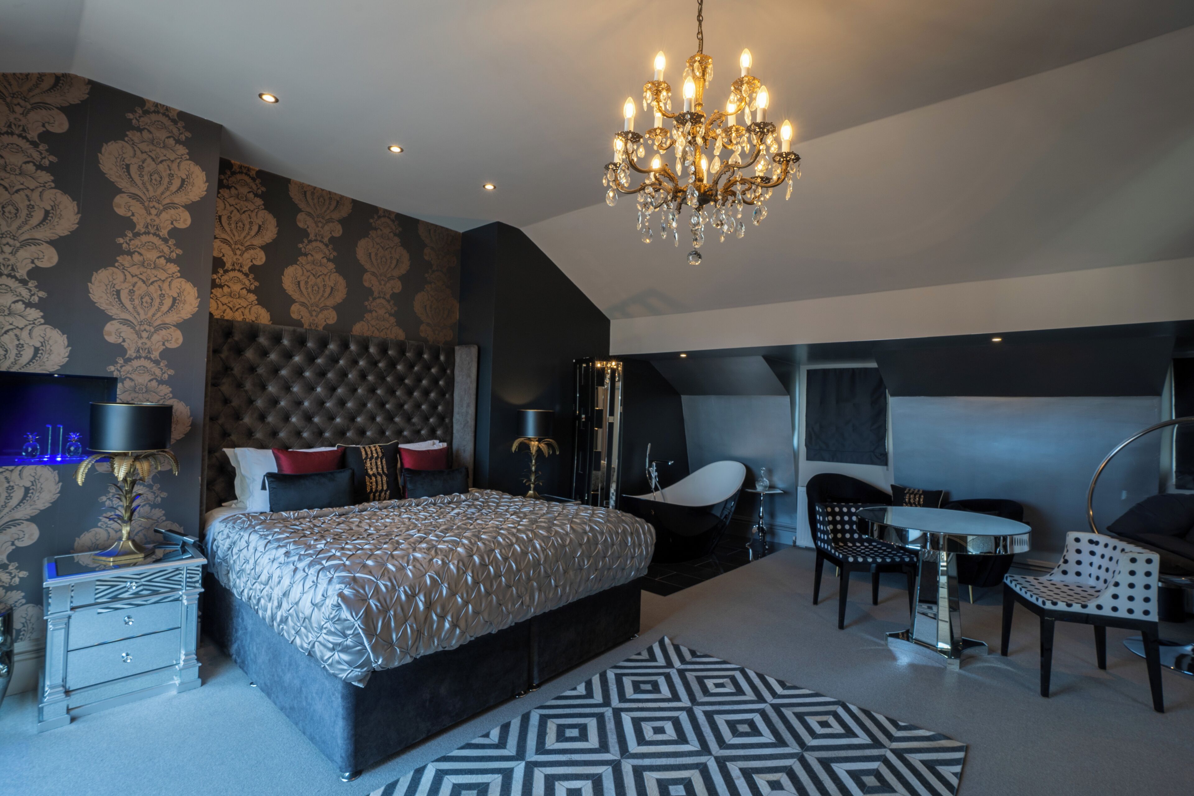 A modern bedroom with a large bed, a chandelier, a vanity, and a seating area with a table and chairs.