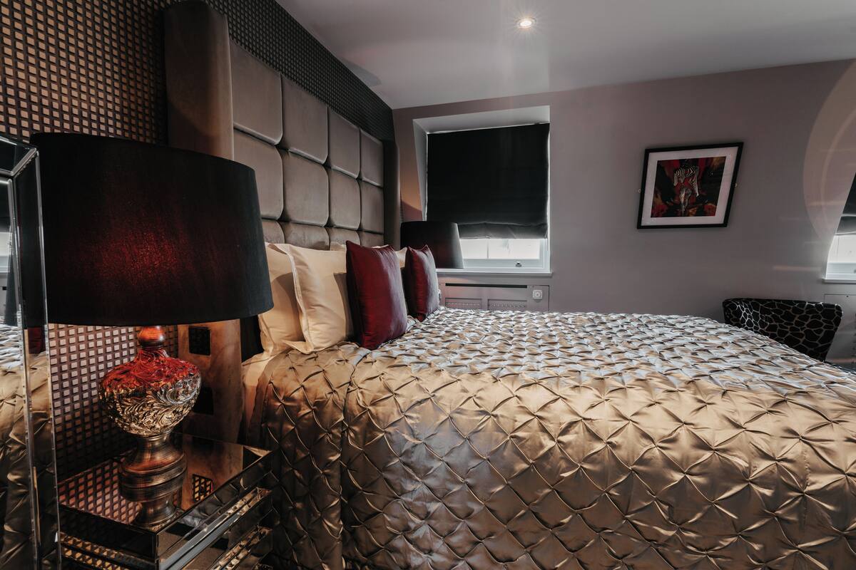 A modern bedroom with a large bed, a headboard with a geometric pattern, a bedside table with a lamp, and a window with a view.