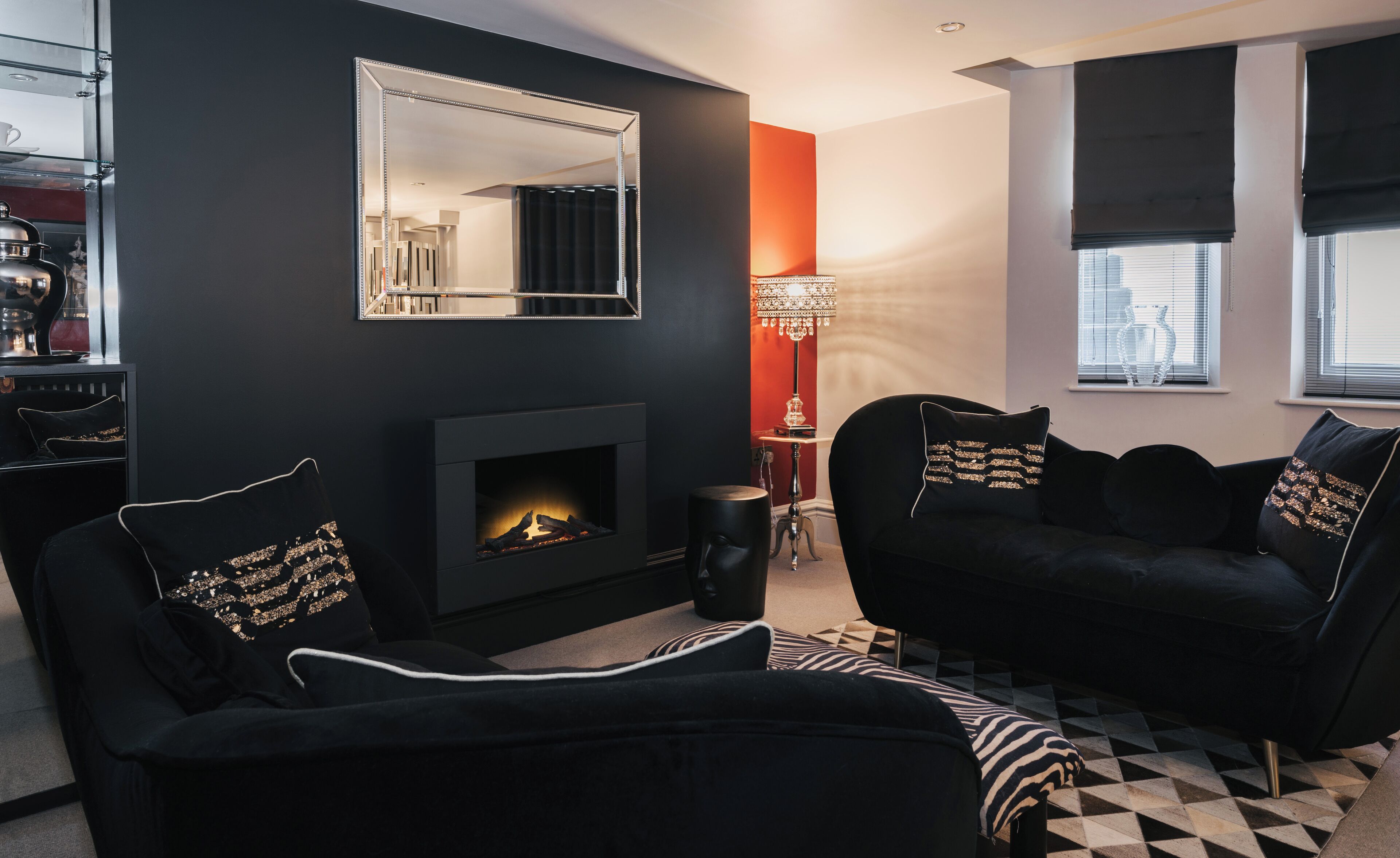 A modern living room with a fireplace, black sofas with gold accents, a large mirror, and a geometric patterned rug.