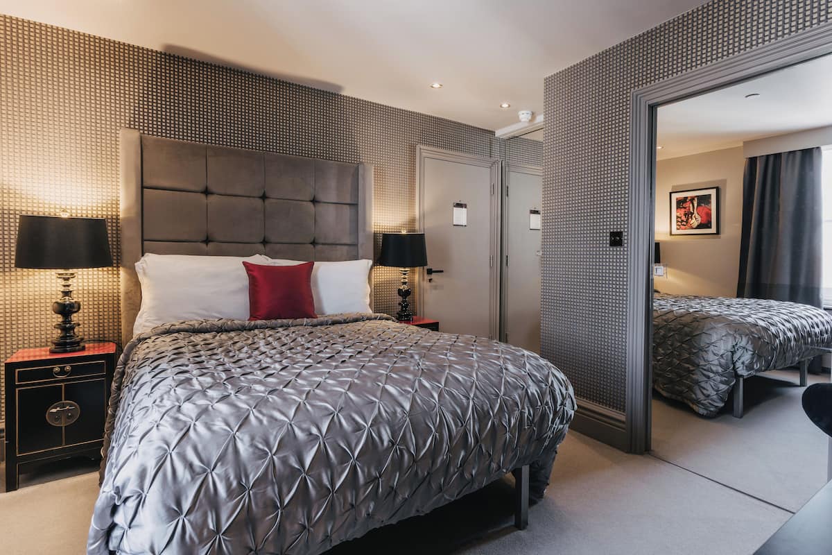 A modern hotel room with a large bed, two bedside tables with lamps, a wardrobe, and a mirror reflecting the room.