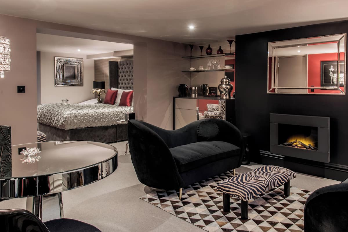 A modern hotel room with a bed, a sofa, a small table, a fireplace, and a mirror.