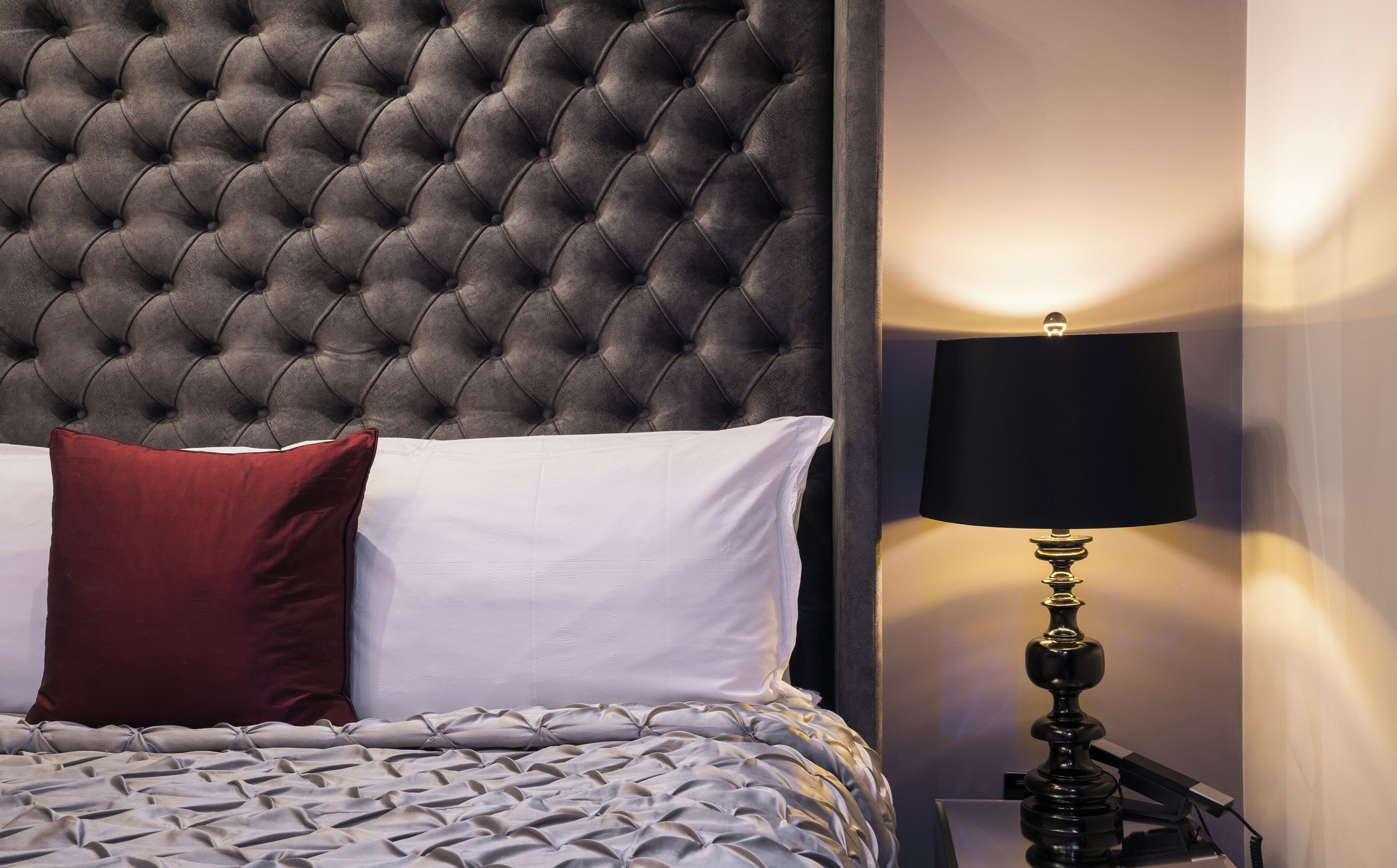 A neatly made bed with a tufted headboard, a red and white pillow, and a lamp on a bedside table.
