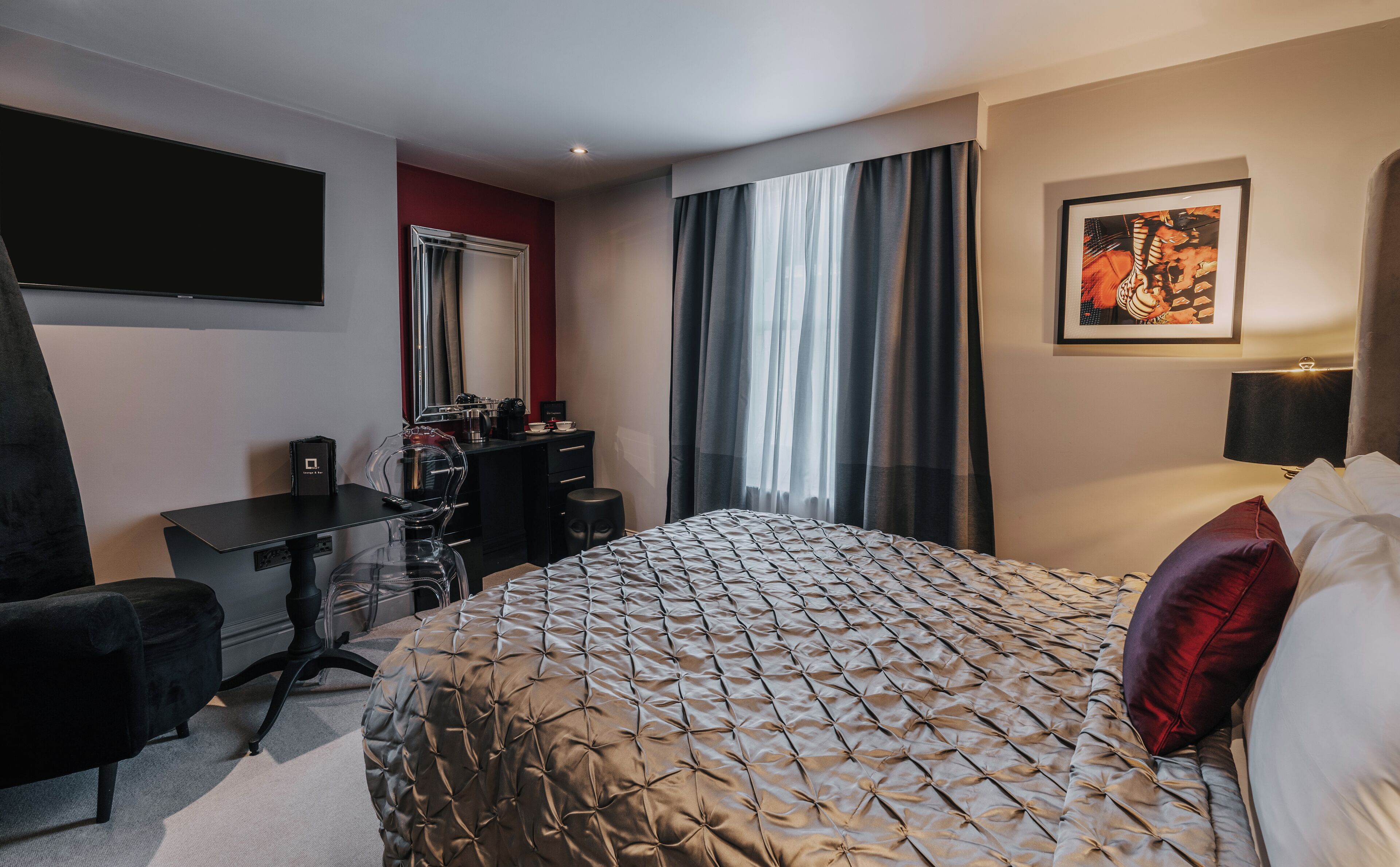A hotel room with a bed, a desk, a chair, a TV, a window with curtains, and a framed picture on the wall.
