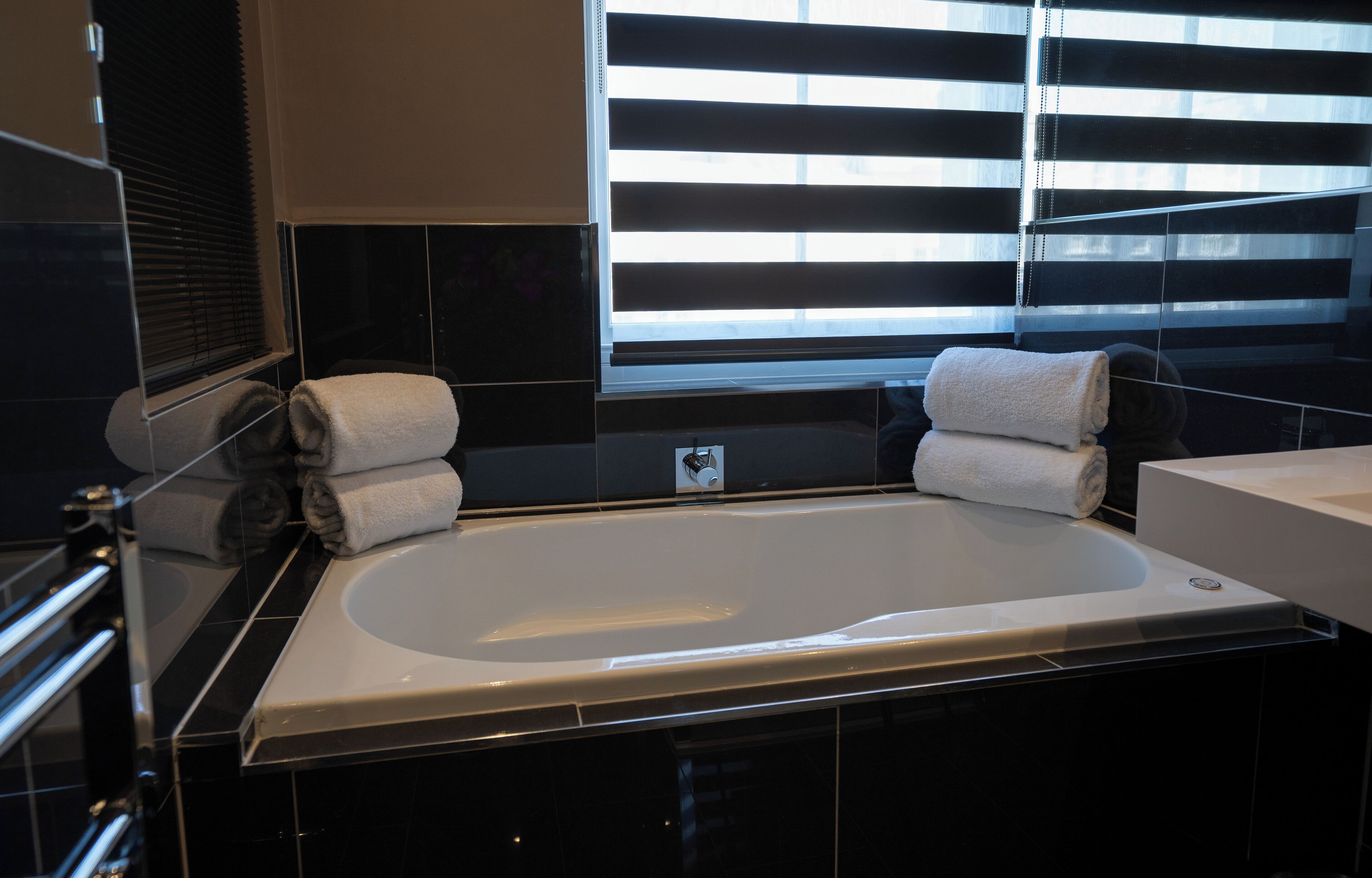 A modern bathroom with a large bathtub, black tiled walls, and a window with blinds.