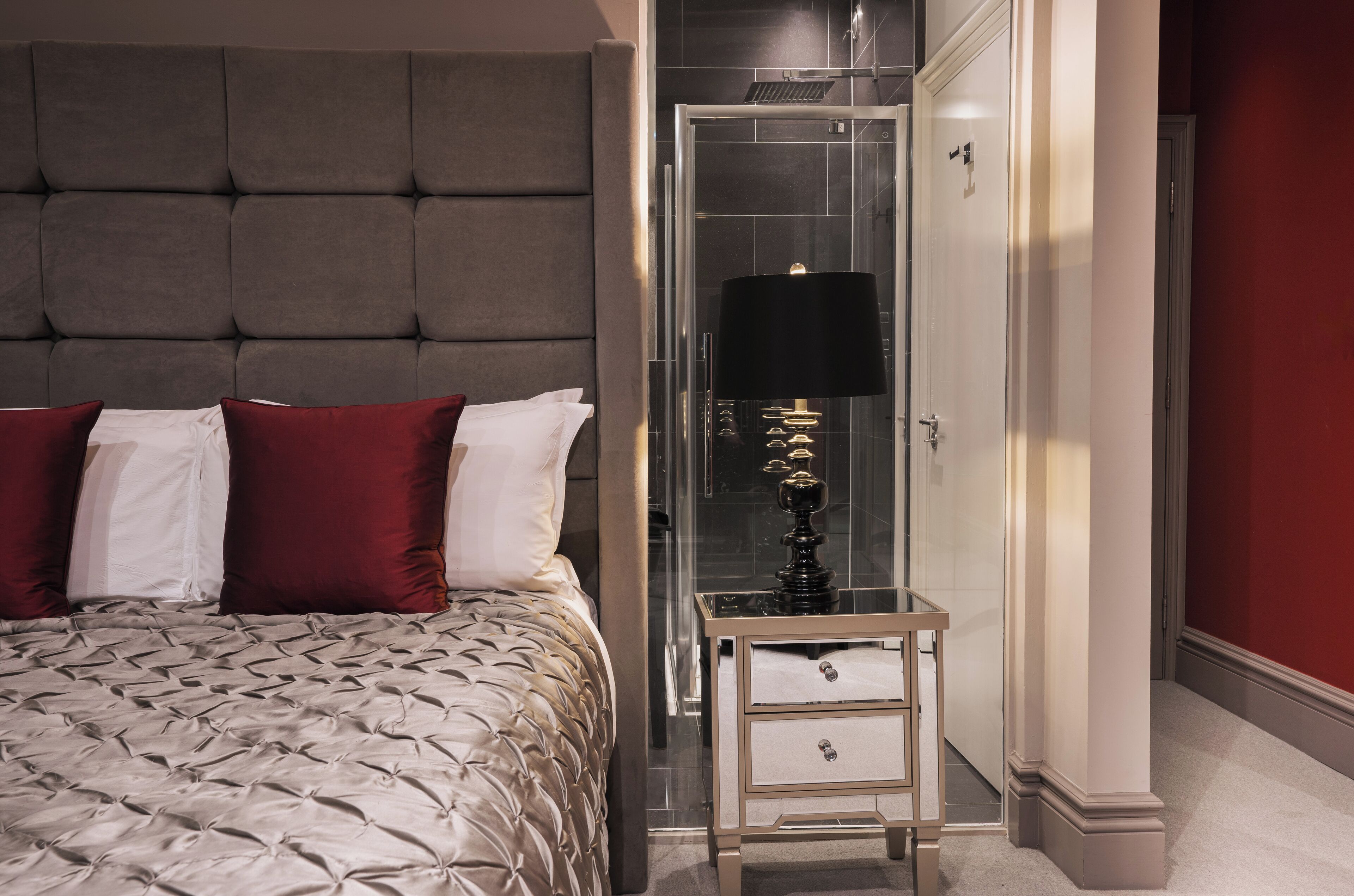 A bedroom with a tufted headboard, a bed with patterned bedding, a bedside table with a lamp, and a glass shower enclosure.