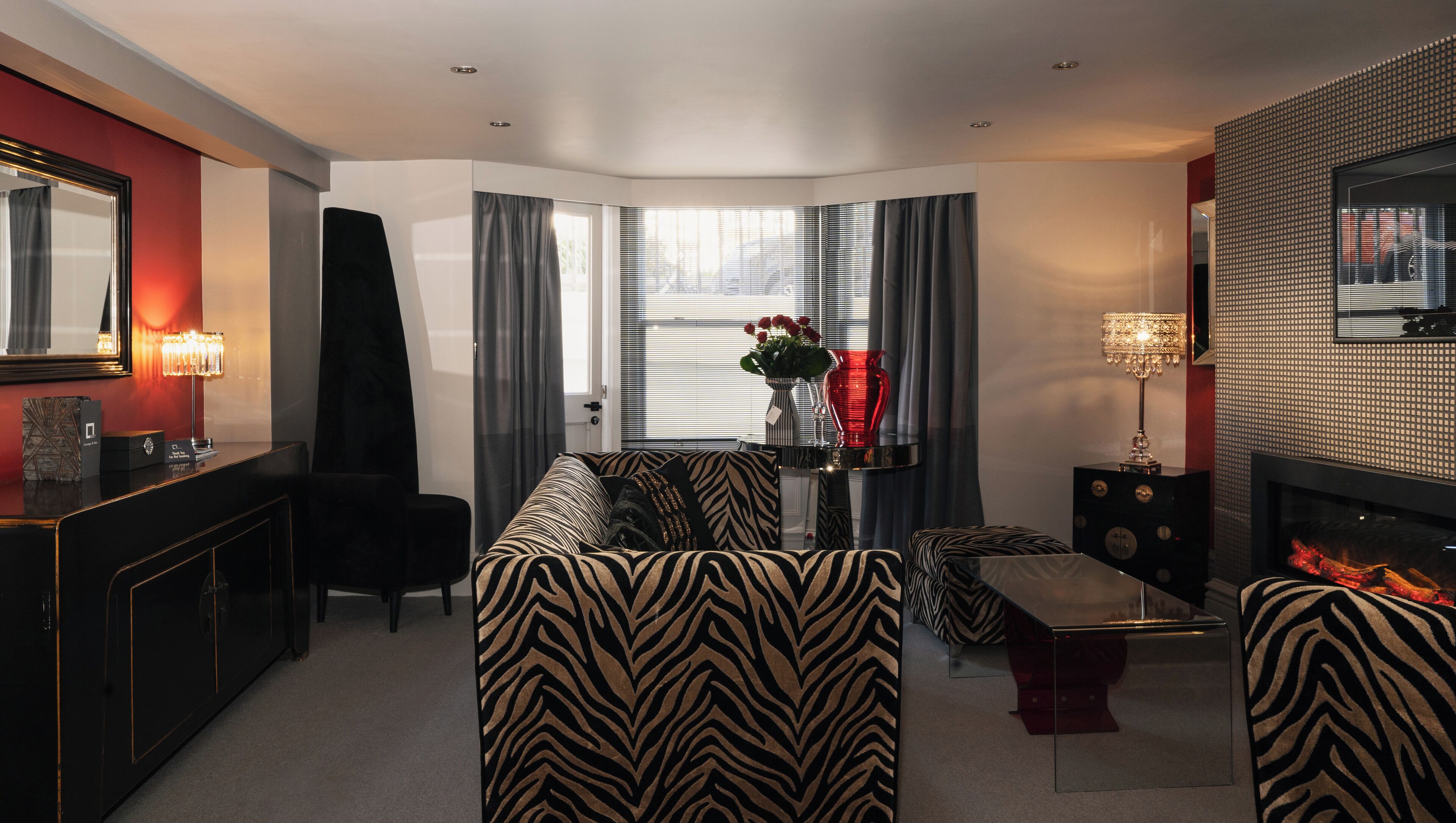 A modern living room with a zebra-patterned sofa, a black coffee table, and a side table with a lamp. There is a fireplace with a red accent and a large mirror on the wall.
