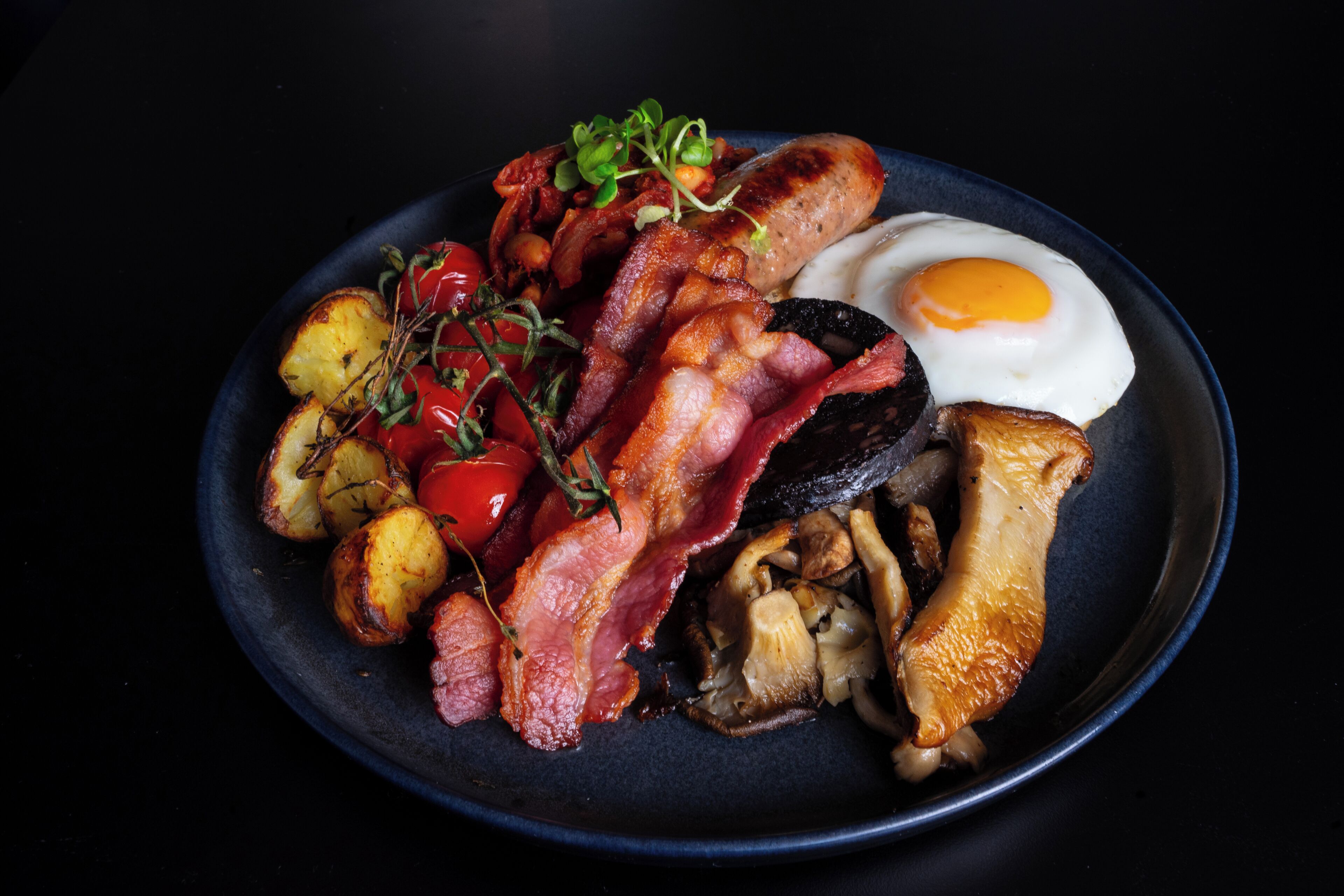 A plate of breakfast food including bacon, eggs, mushrooms, tomatoes, and roasted potatoes.
