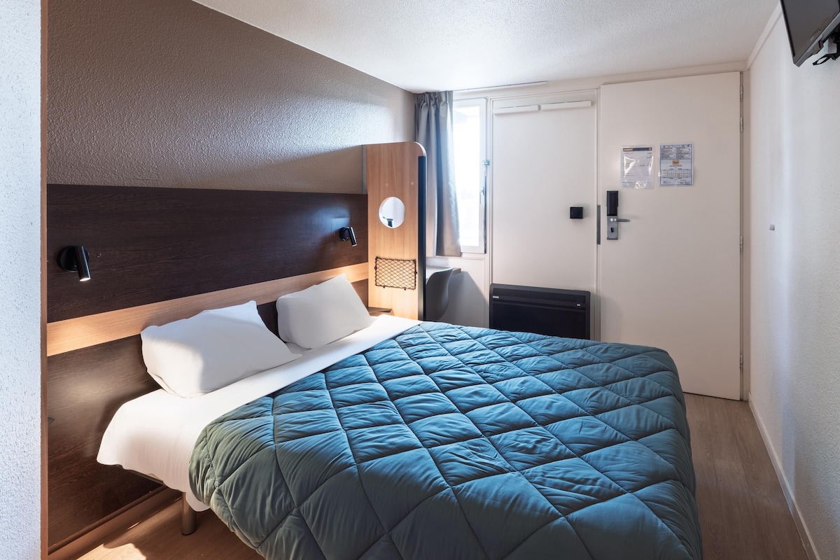 A hotel room with a bed, a nightstand, a wall-mounted lamp, and a door with a handle.
