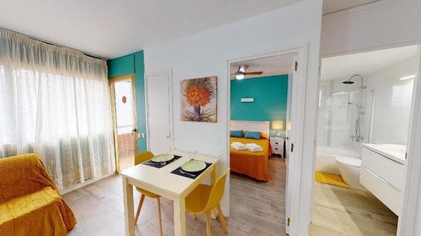 Habitación de hotel con zona de comedor, cama con ropa de cama naranja, zona de ducha y un cuadro en la pared.