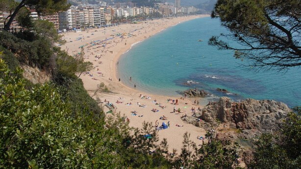 A coastal beach with a rocky shoreline, a sandy beach area, and a row of buildings in the background. - Eldorado Apartamentos Turisticos (Lloret de Mar)