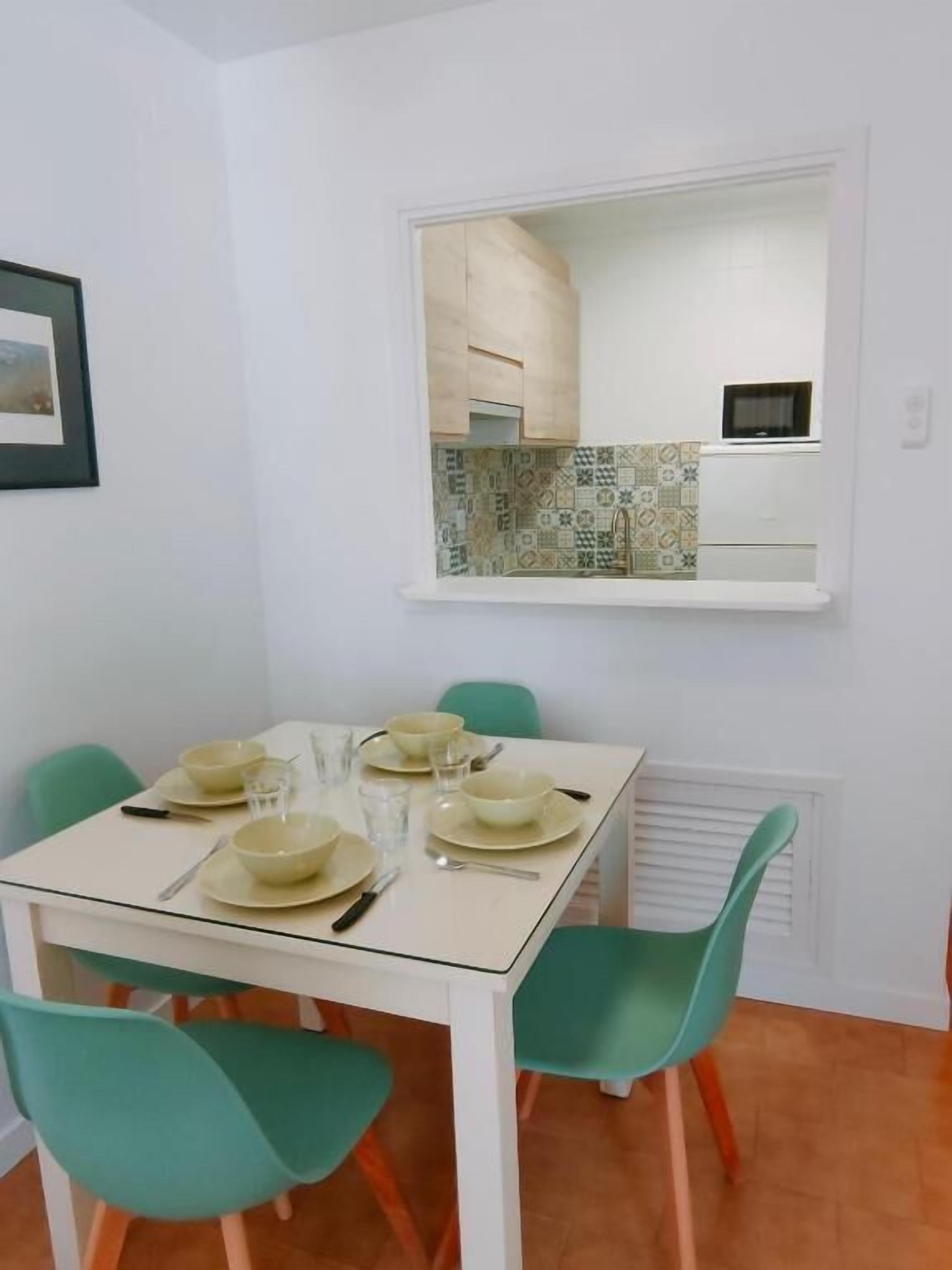 Un comedor con una mesa dispuesta para cuatro, cuatro sillas verdes y una cocina visible a través de una ventana.