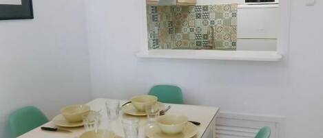 Un comedor con una mesa dispuesta para cuatro, cuatro sillas verdes y una cocina visible a través de una ventana.