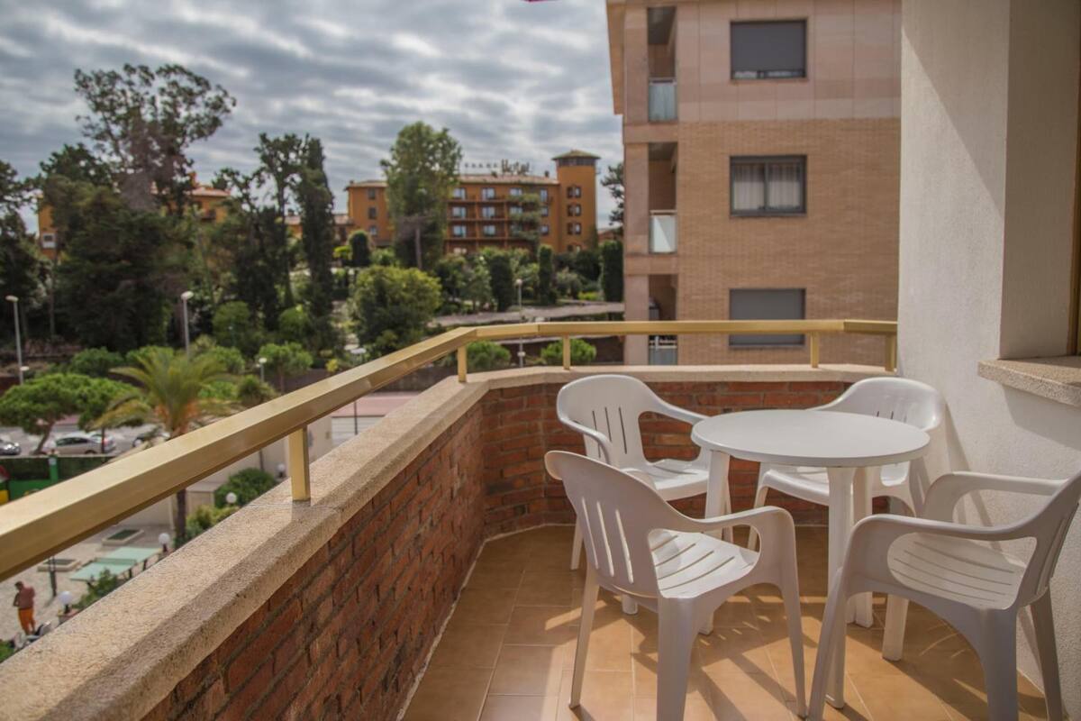 Un balcón con sillas de plástico blanco y una pequeña mesa redonda, con vista a un edificio de ladrillo y una calle con coches y árboles.