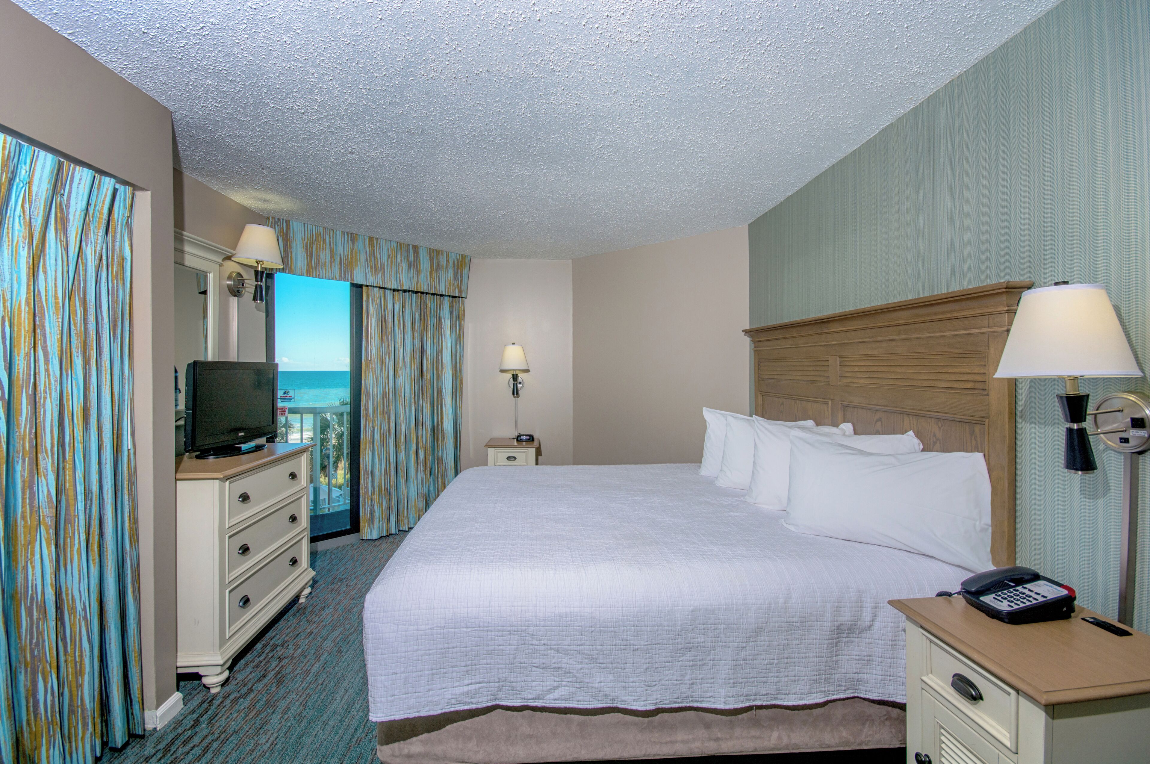 A hotel room with a bed, a television on a dresser, a nightstand with a lamp, and a phone on a desk.
