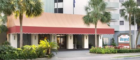 A hotel entrance with a red awning, palm trees, and a sign indicating 'The Strand Boutique Resort'.