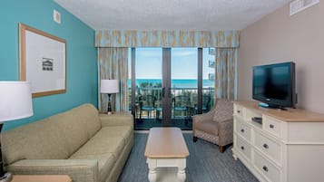 A hotel room with a sofa, a coffee table, a TV on a dresser, and a balcony with a view of the ocean.