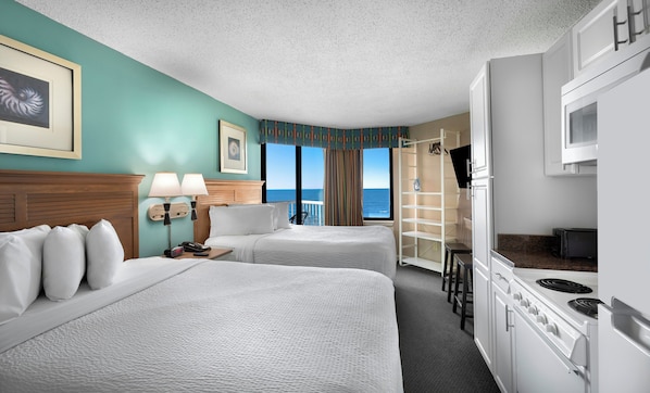A hotel room with two beds, a kitchenette, a TV, and a view of the ocean.