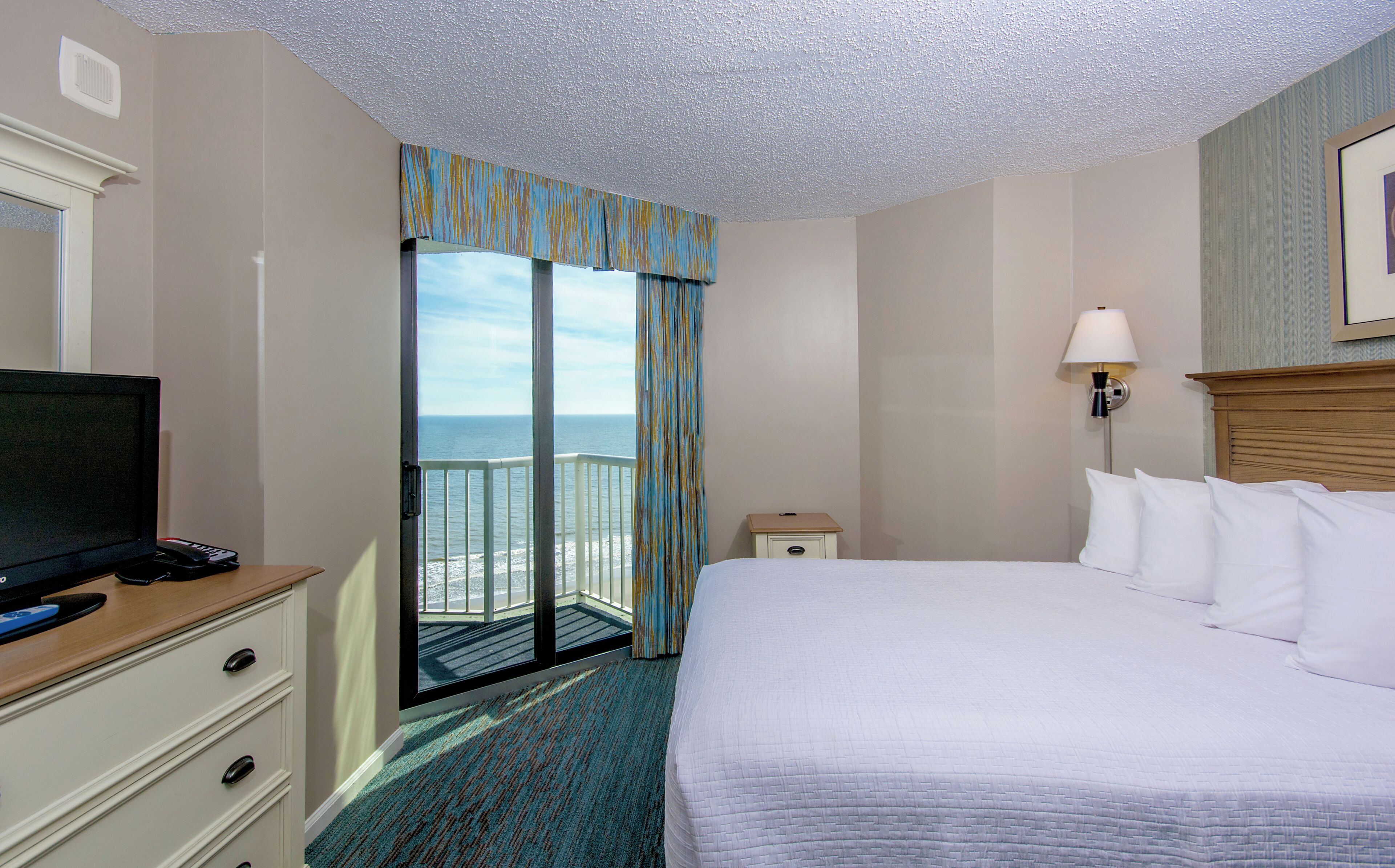 A hotel room with a bed, a television, a desk with a lamp, and a view of the ocean.
