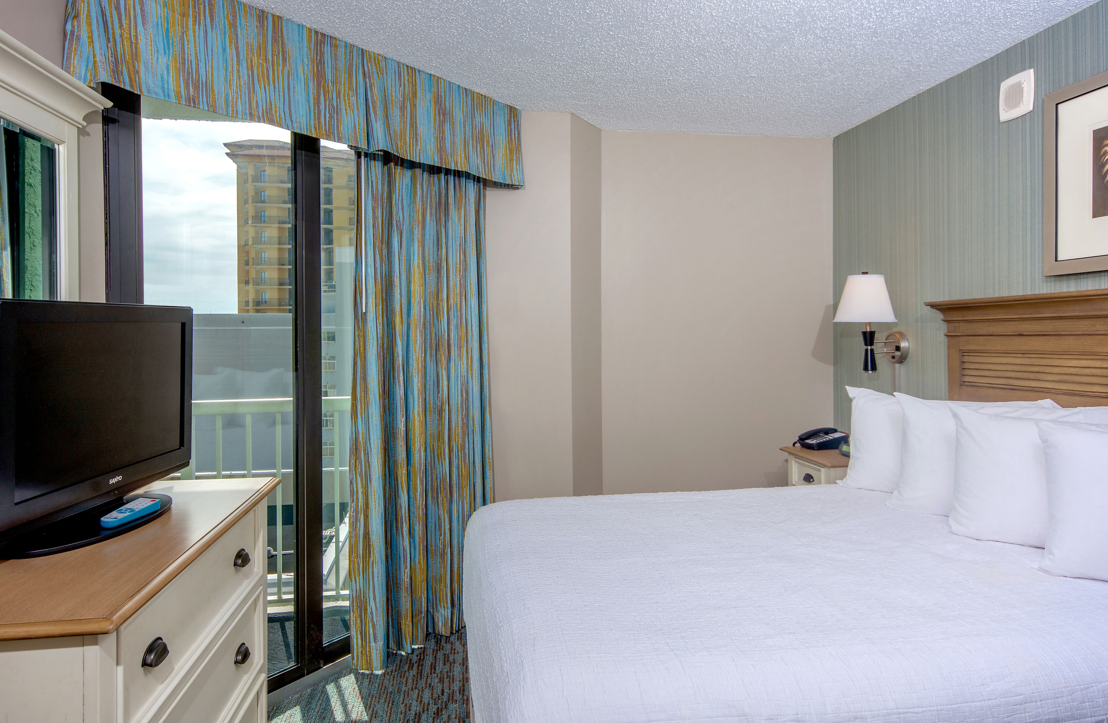 A hotel room with a large bed, a TV on a dresser, a nightstand with a lamp, and a view of a building outside.
