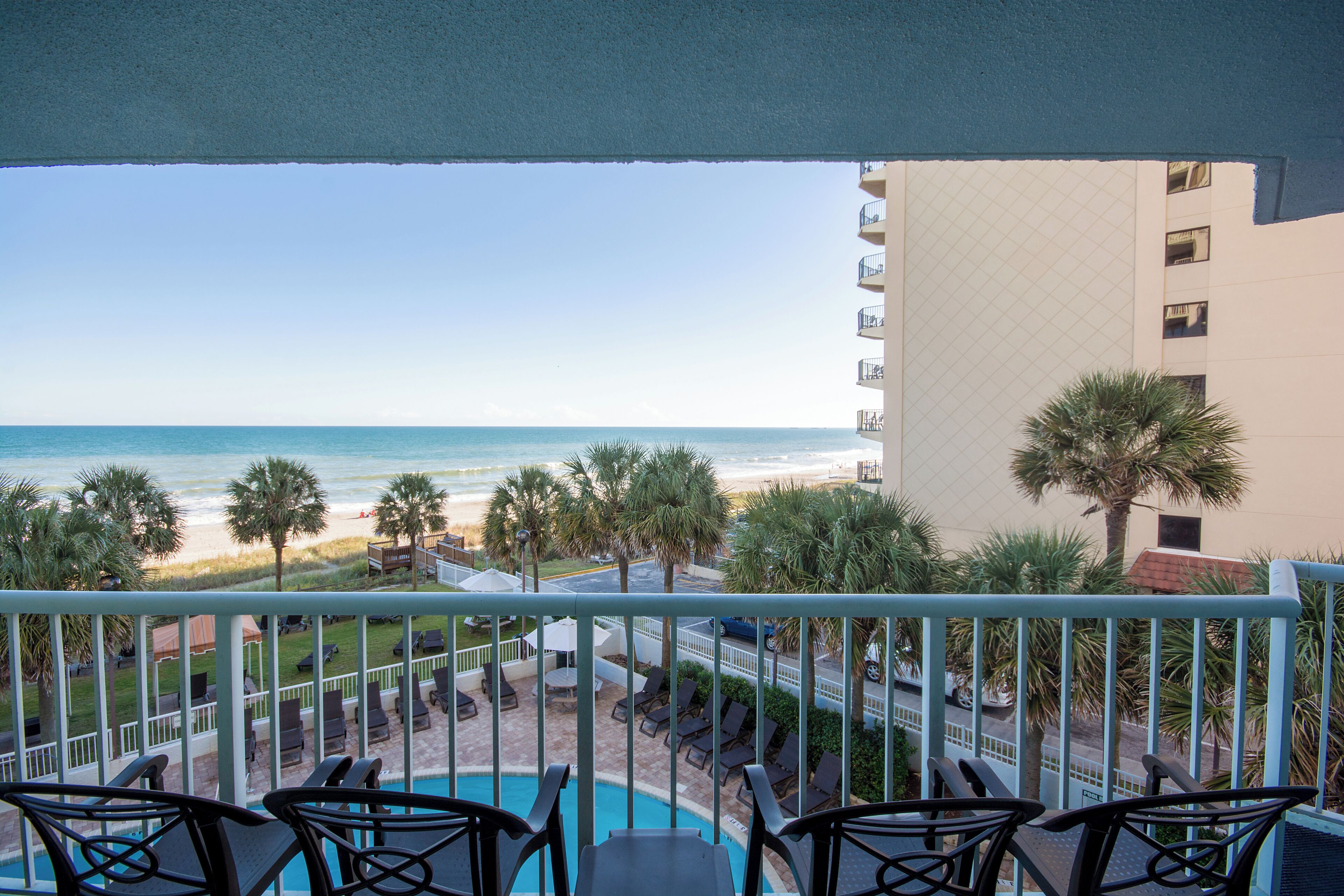 A balcony with a view of the beach, poolside chairs, and a building with balconies.