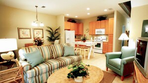 A living room with a striped sofa, a round coffee table with a plant, a green armchair, and a kitchen in the background. - Coral Cay Resort by Sky Hotels & Resorts (Kissimmee)