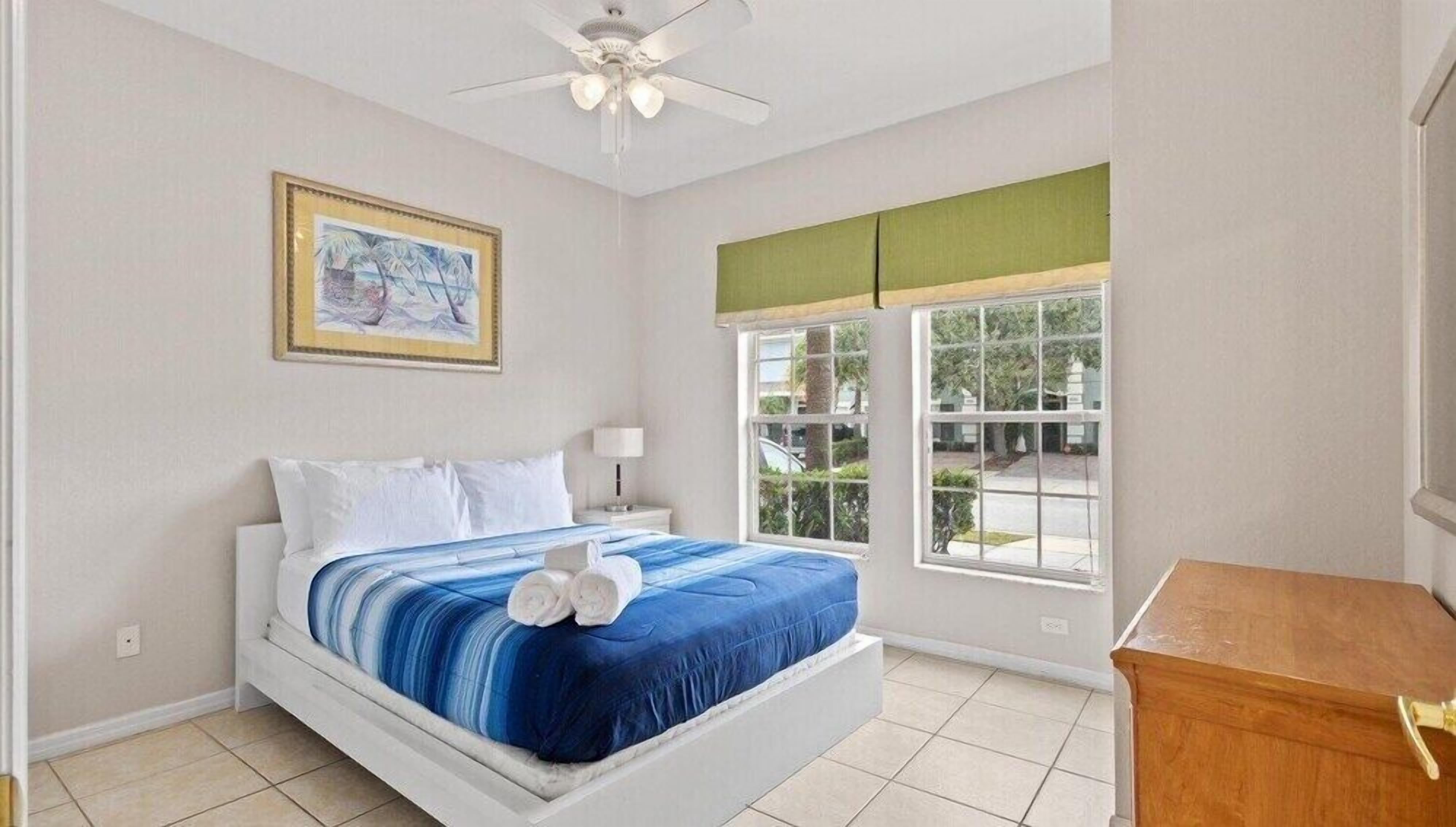 A bedroom with a bed, a nightstand, a ceiling fan, a window with a view, and a framed picture on the wall.