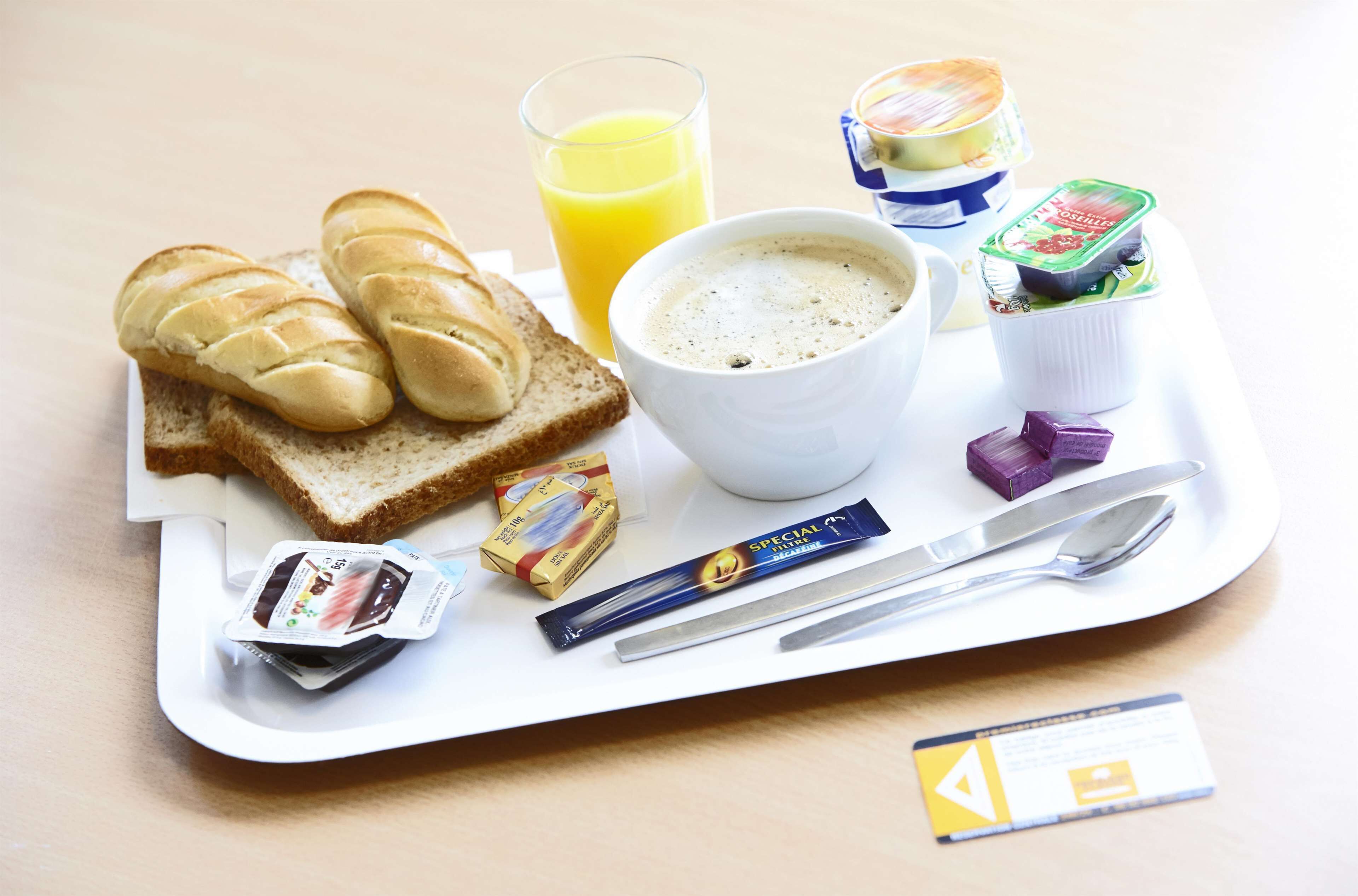 A breakfast tray with toast, a cup of coffee, a glass of orange juice, a container of yogurt, a chocolate bar, a packet of instant coffee, and a spoon and fork.