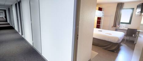 A hotel corridor with a bed, desk, and window.