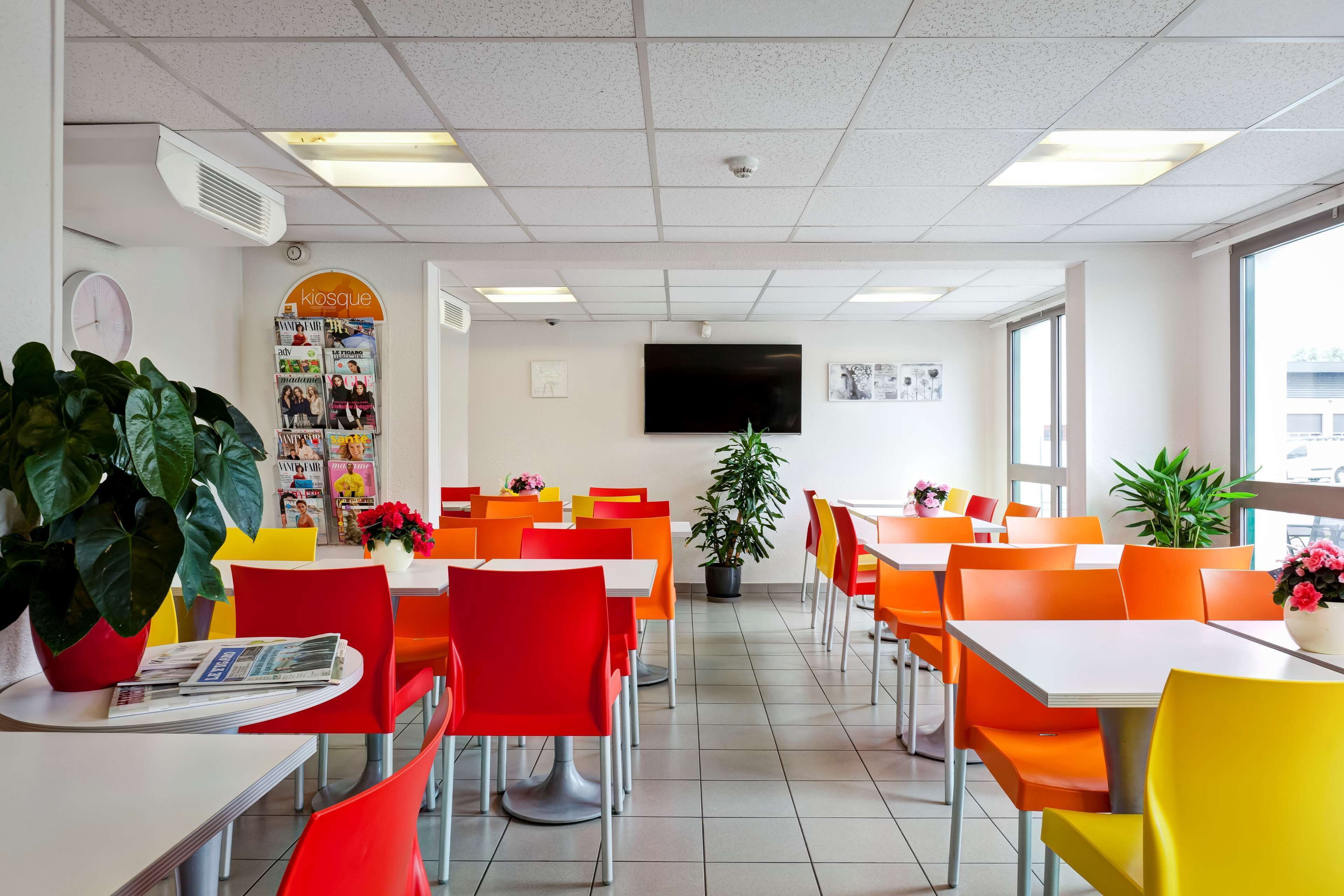 A brightly lit cafeteria with orange and yellow chairs, white tables, and potted plants.