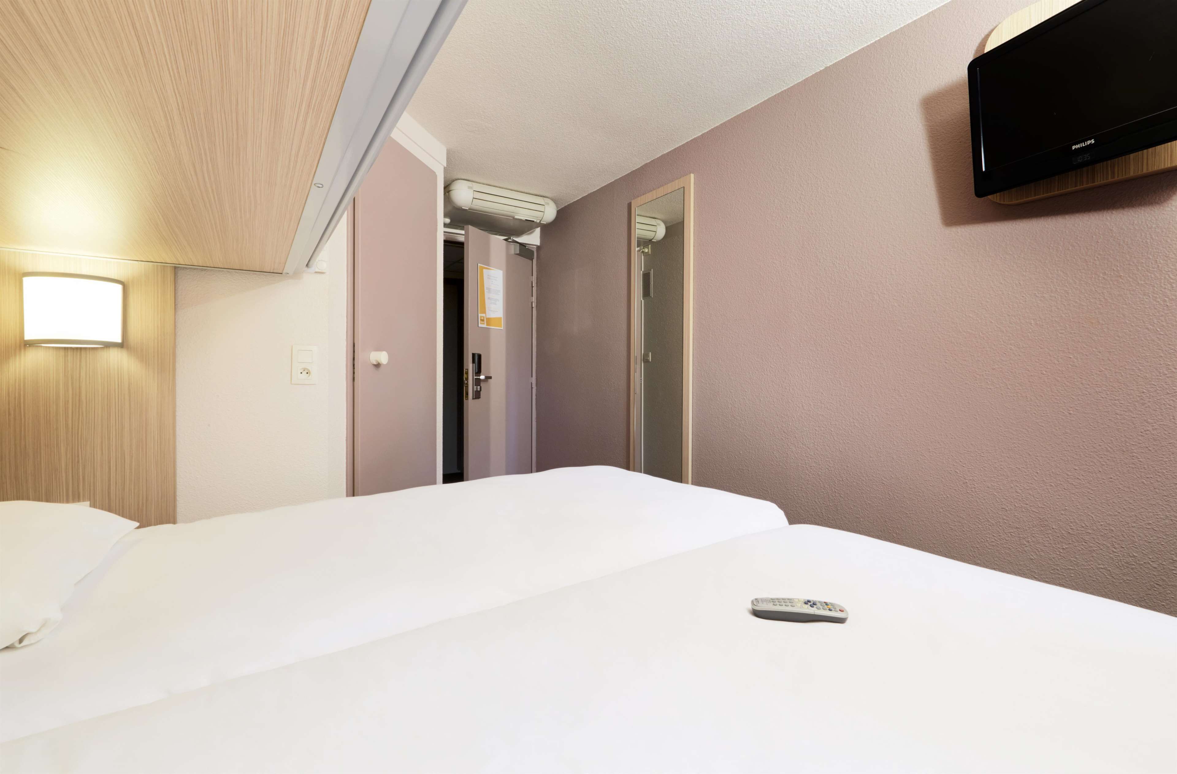 A hotel room with a bed, a remote control on the bed, a wall-mounted TV, and a door leading to another room.