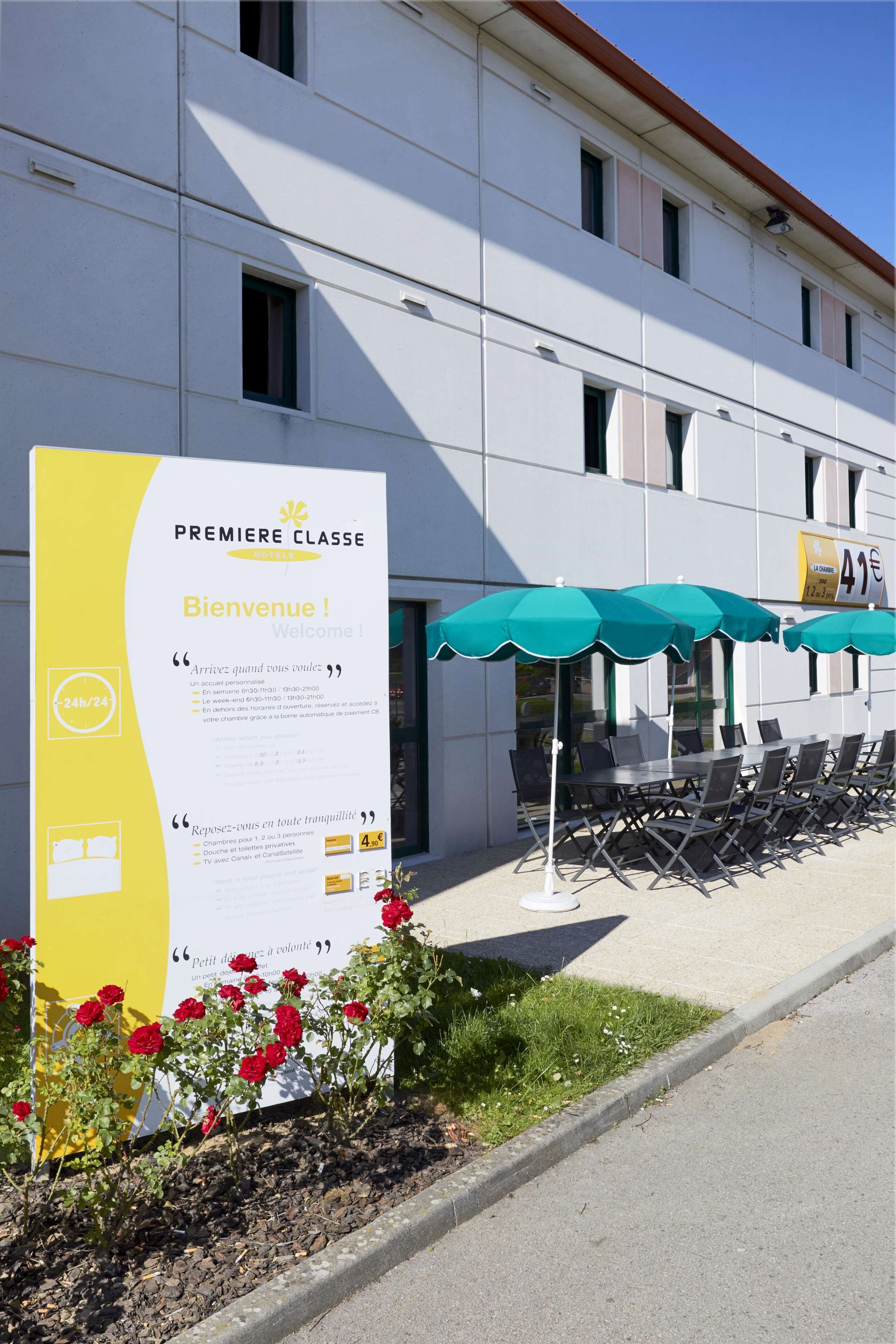 A hotel entrance with a large welcome sign, outdoor seating with umbrellas, and a flower bed.