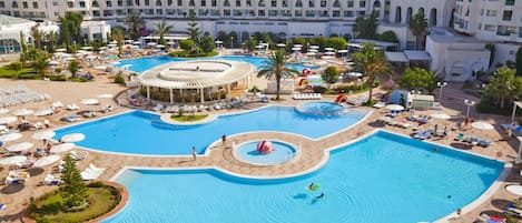 Une grande piscine dotée de plusieurs zones et d’une fontaine centrale, entourée de chaises longues et de parasols.