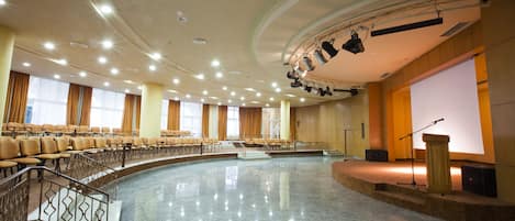Un amplio salón bien iluminado con asientos escalonados, un escenario con un atril y una pantalla grande.