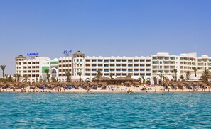 A large hotel with multiple balconies, a beach area with umbrellas, and palm trees.