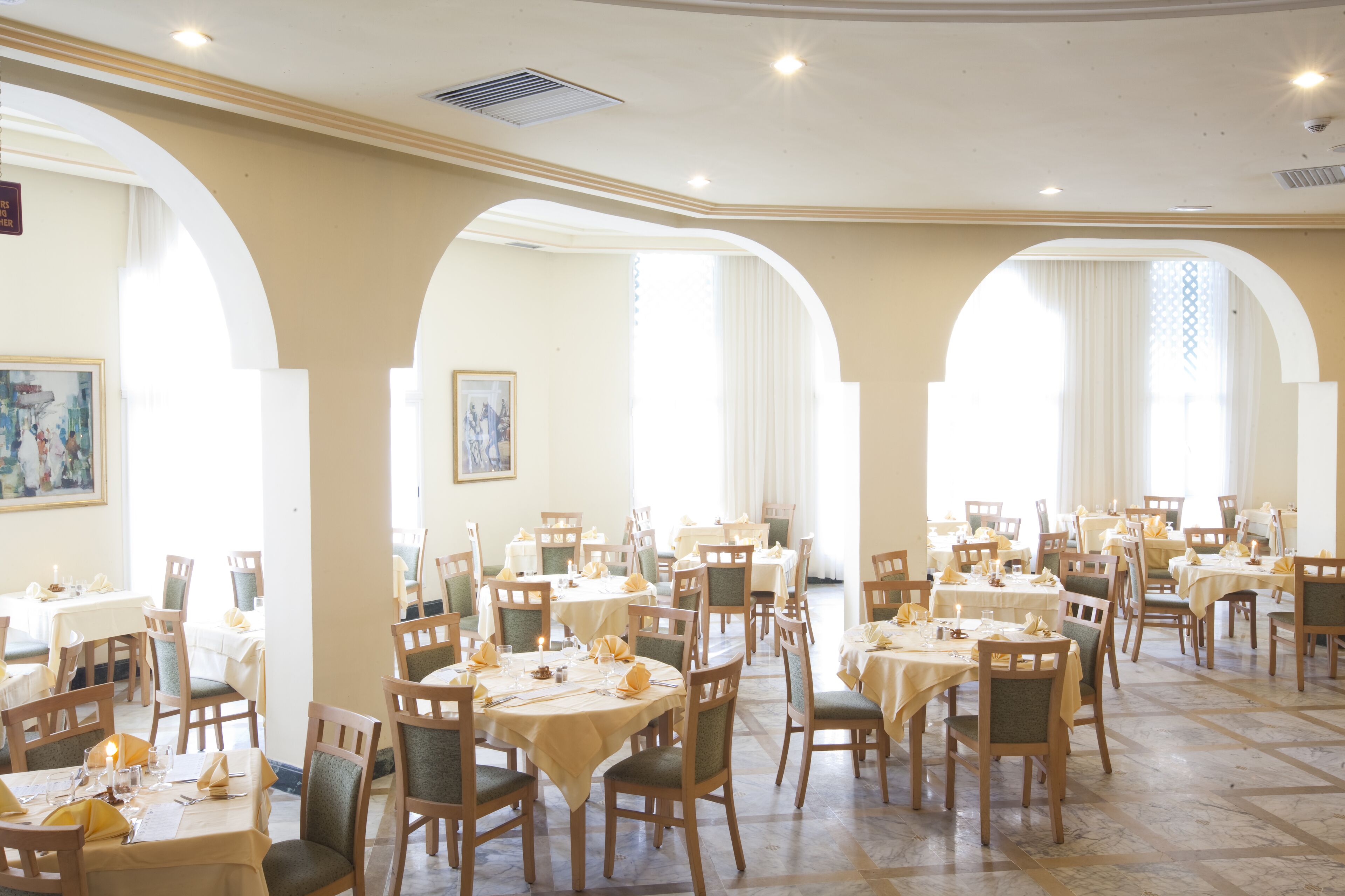 A dining area with multiple tables and chairs, arranged in a spacious layout with large windows and arches.