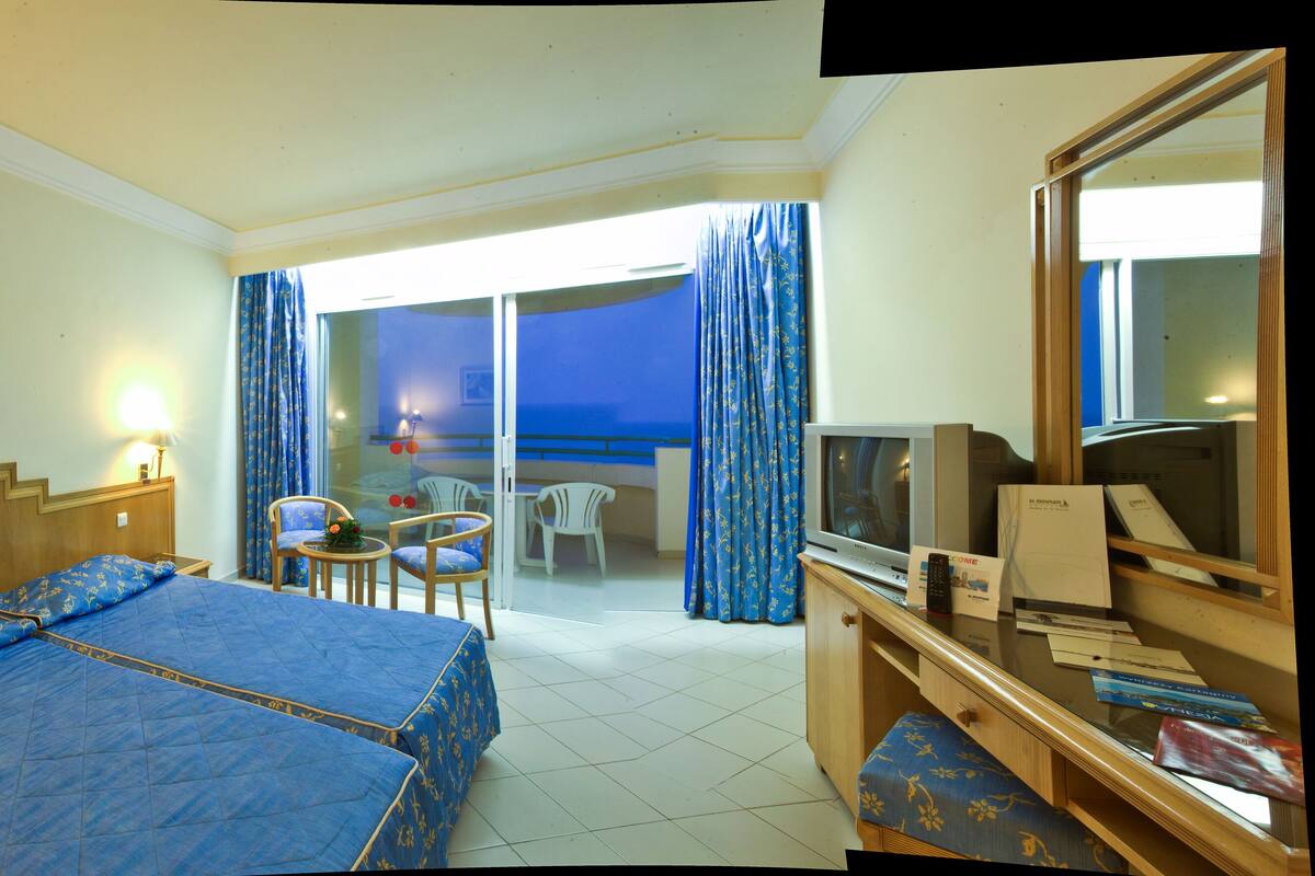 Een hotelkamer met een bed, een bureau met een computer, een televisie en een balkon met uitzicht.