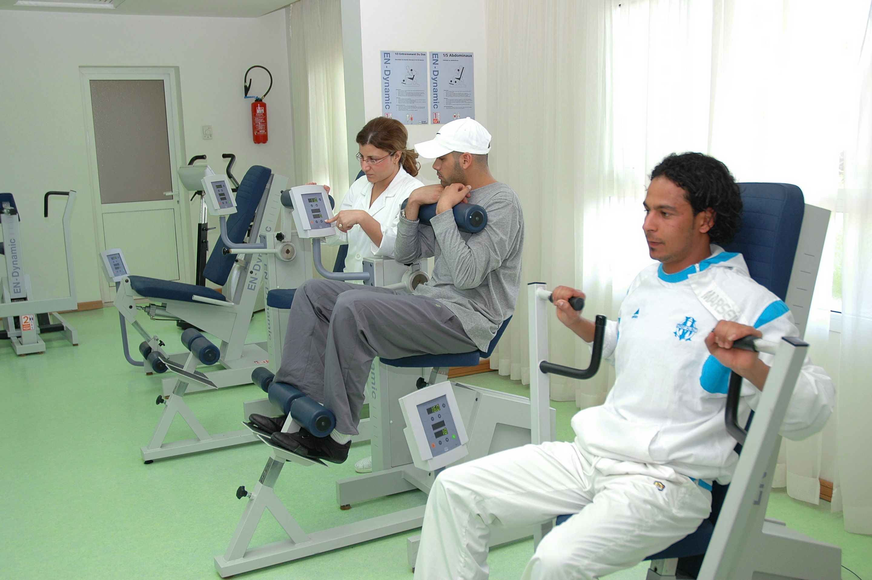 A medical examination room with patients seated on examination tables, a fire extinguisher on the wall, and informational posters.