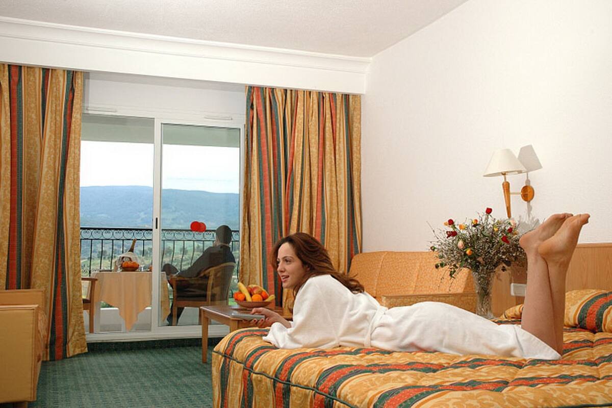Une chambre d’hôtel avec un lit, une table sur laquelle se trouve un panier de fruits, une chaise, un balcon avec vue et un vase rempli de fleurs.